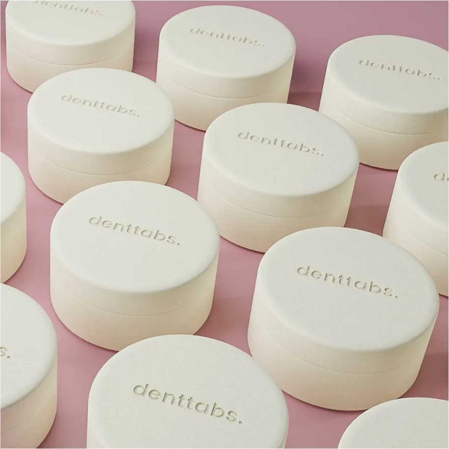 Reihen cremefarbener Denttabs-Dosen mit geprägtem Logo auf rosa Grund