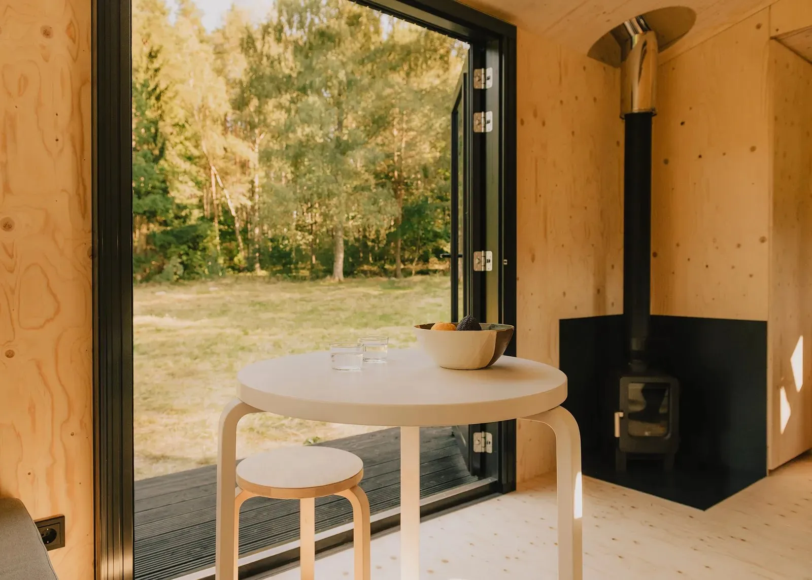 Innenraum einer StayRaus-Kabine mit Ofen, Tisch und Blick in den Wald